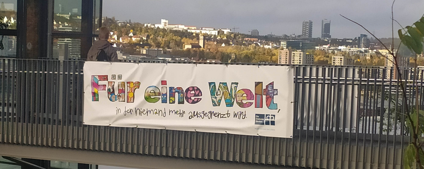 Banner mit Aufschrift ‚Für eine Welt, in der niemand mehr ausgegrenzt wird‘ am Geländer mit Blick auf die Stadt und moderne Hochhäuser im Hintergrund.